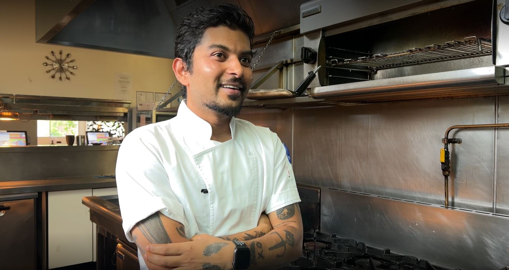 A man with black hair in a white chef outfit smiles in front of a stainless steel kitchen.