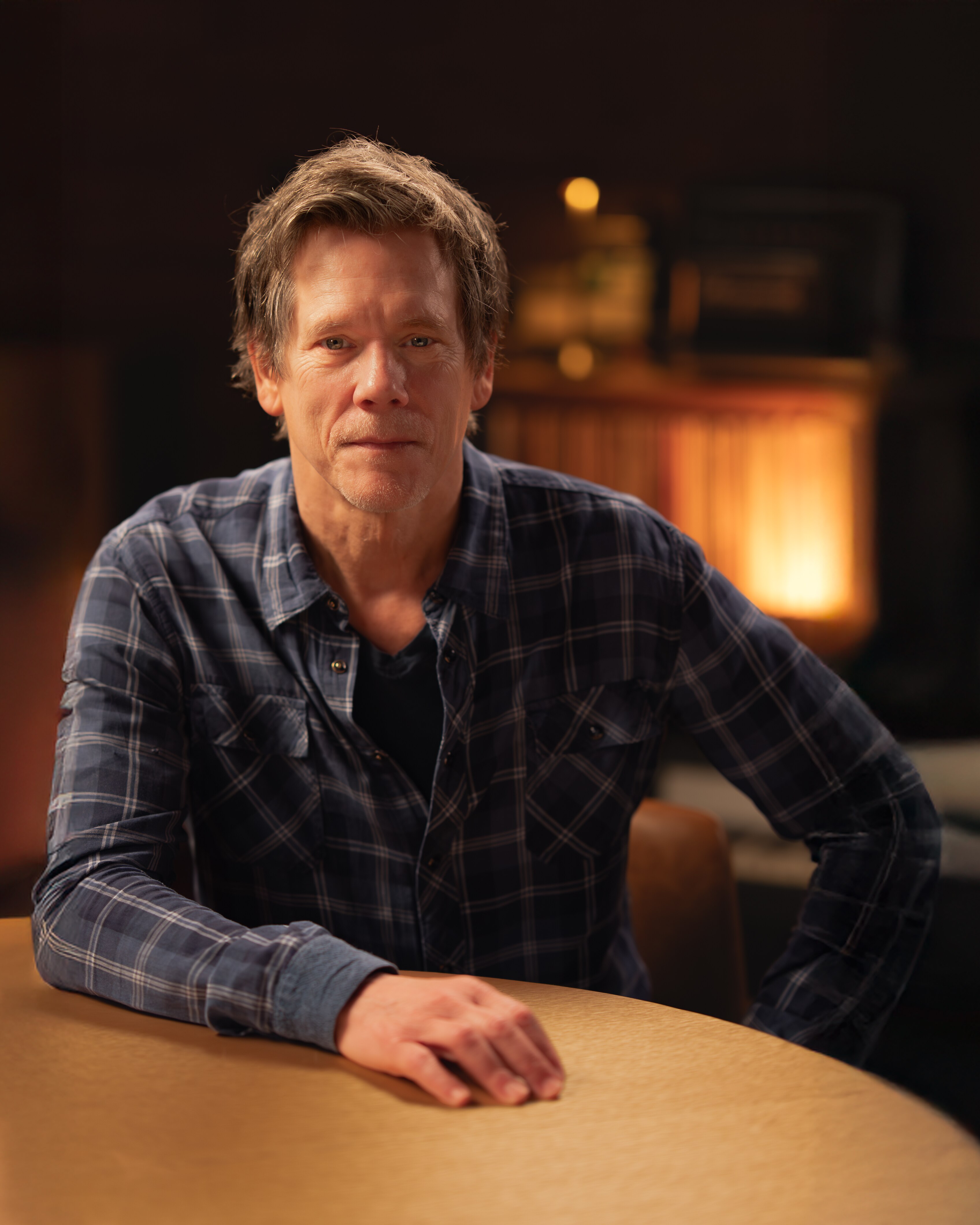 Kevin Bacon in a blue check long-sleeve collared shirt sits in a New York recording studio