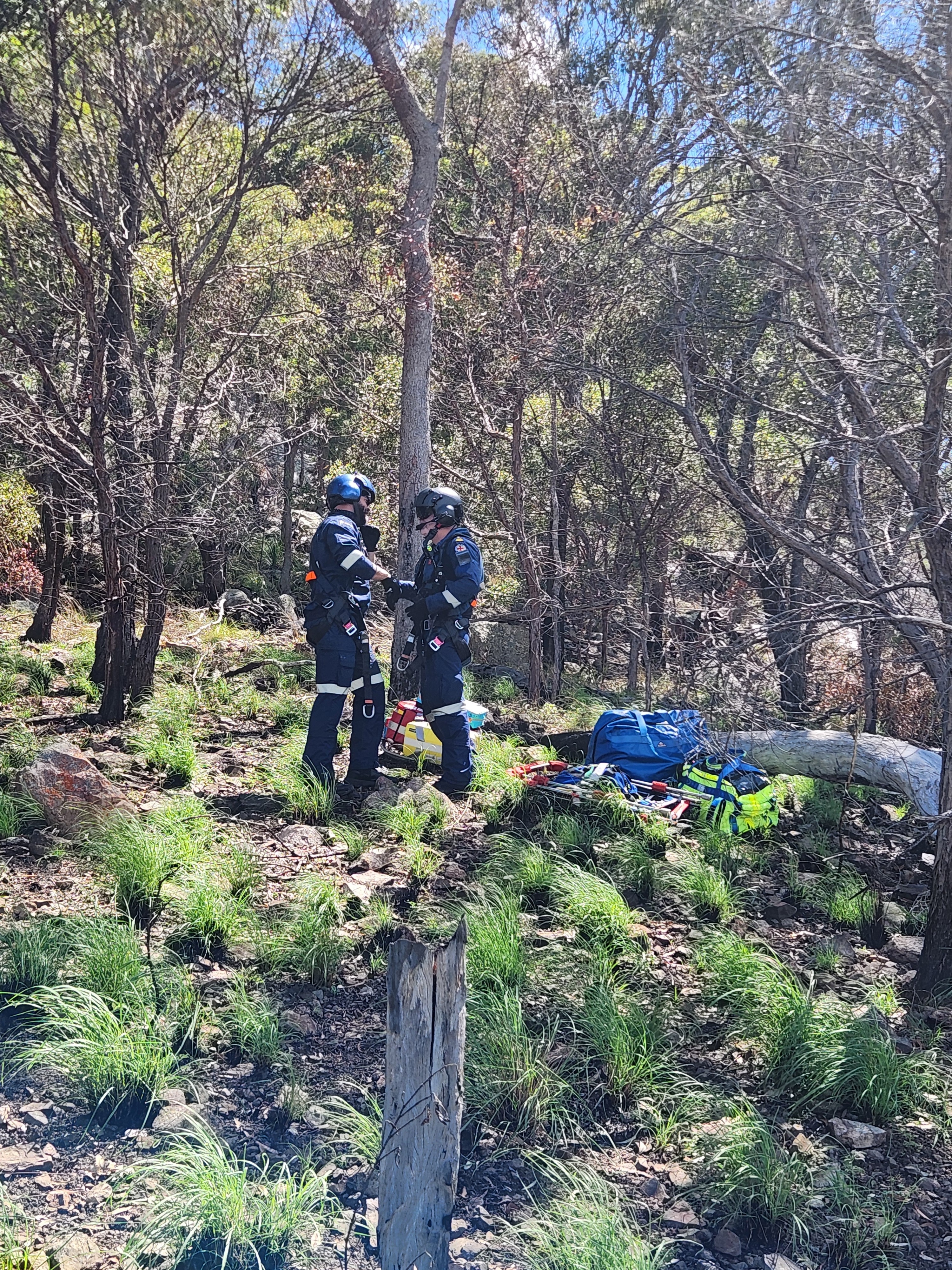 Dois homens com trajes de voo e capacetes em um matagal com equipamentos de resgate ao lado deles.