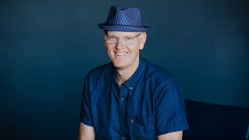 A man smiling to camera in front of a blue backdrop, blue shirt and blue straw hat.