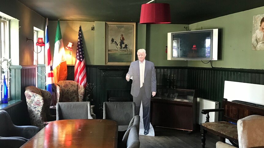 A cardboard cutout of US President Donald Trump in a dim green-painted room with chairs, table, TV and a US flag