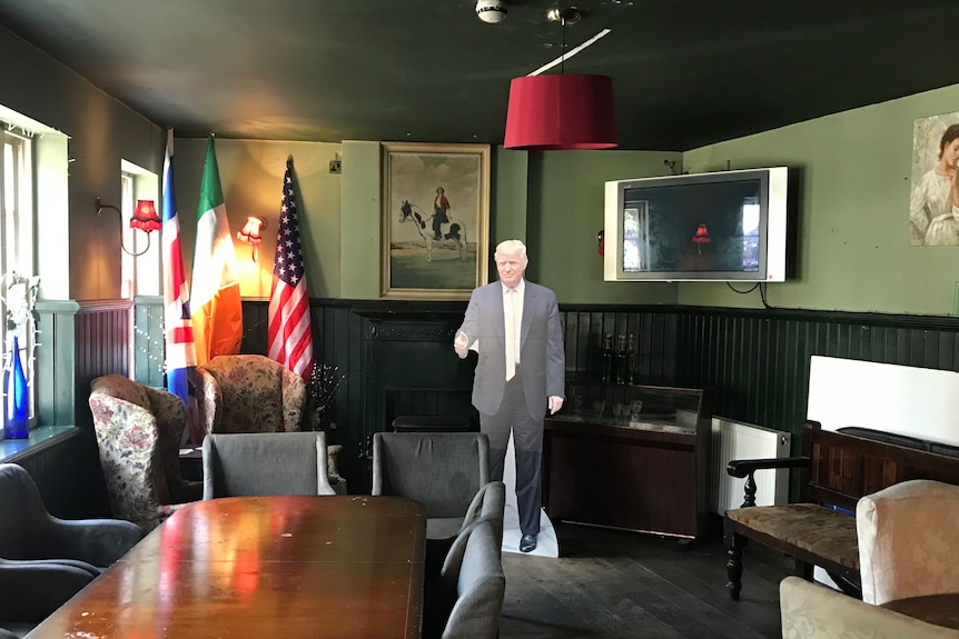 A cardboard cutout of US President Donald Trump in a dim green-painted room with chairs, table, TV and a US flag