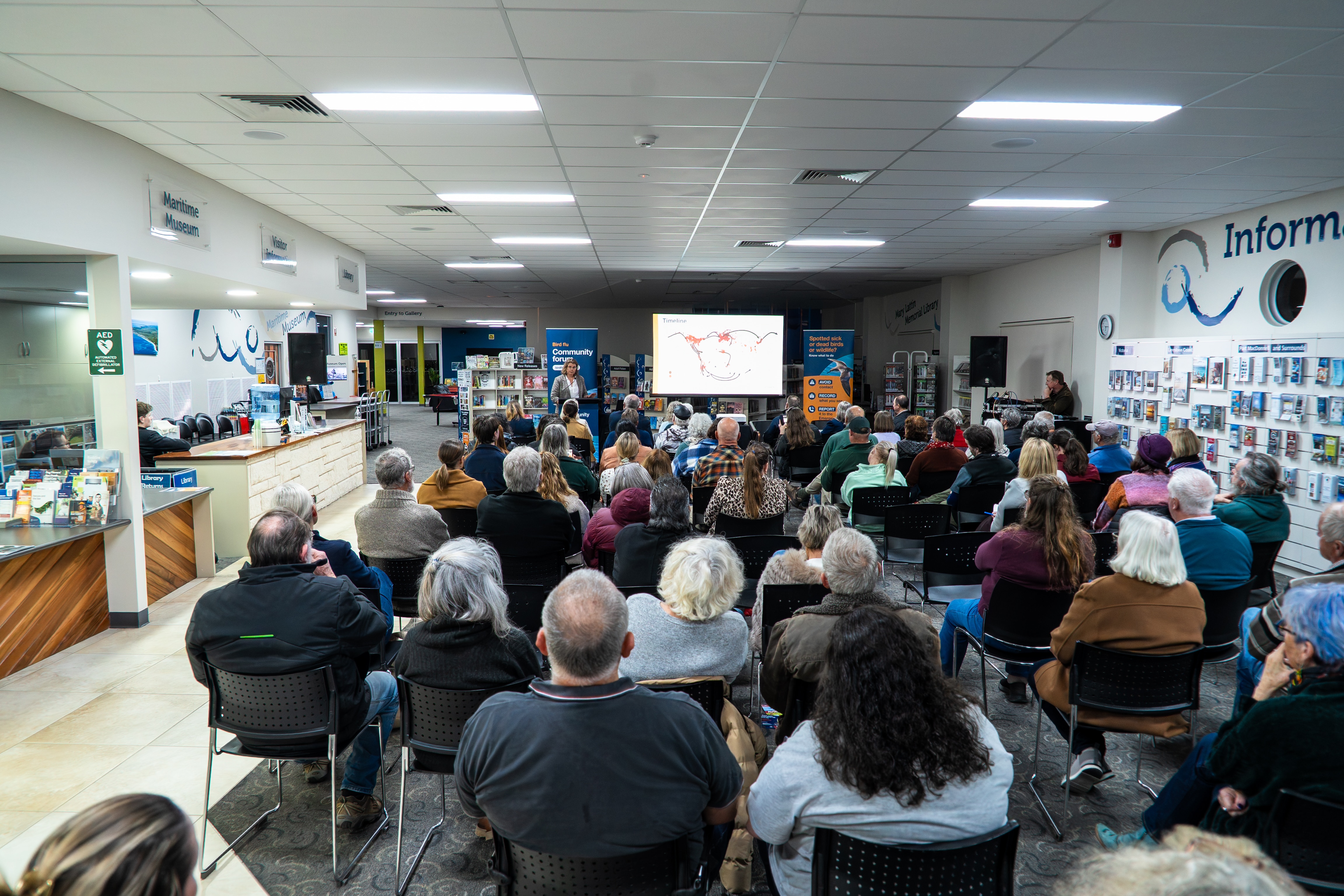 A group of adults sit in plastic chairs as a state government representative goes through slides inside a community centre.