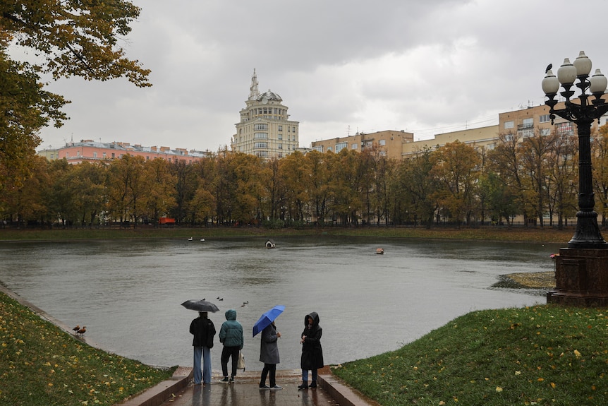 Four women in coats holding umbrellas stand next to a large pond in Moscow.