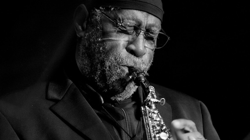 A monochrome shot of Bennie Maupin playing his soprano sax; he's wearing glasses and a beanie