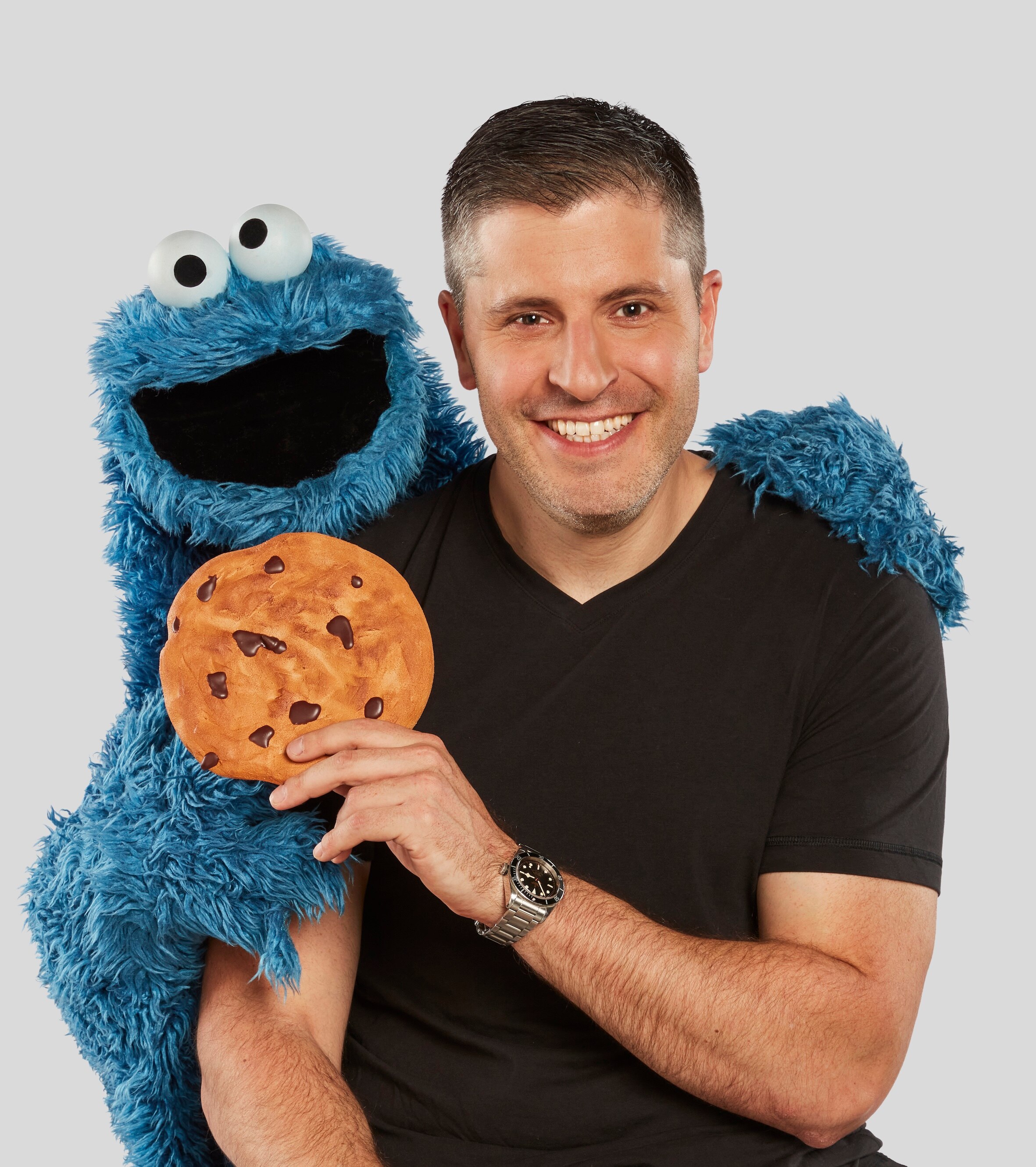 A smiling man in a black shirt arm in arm with a blue monster holding a cookie.
