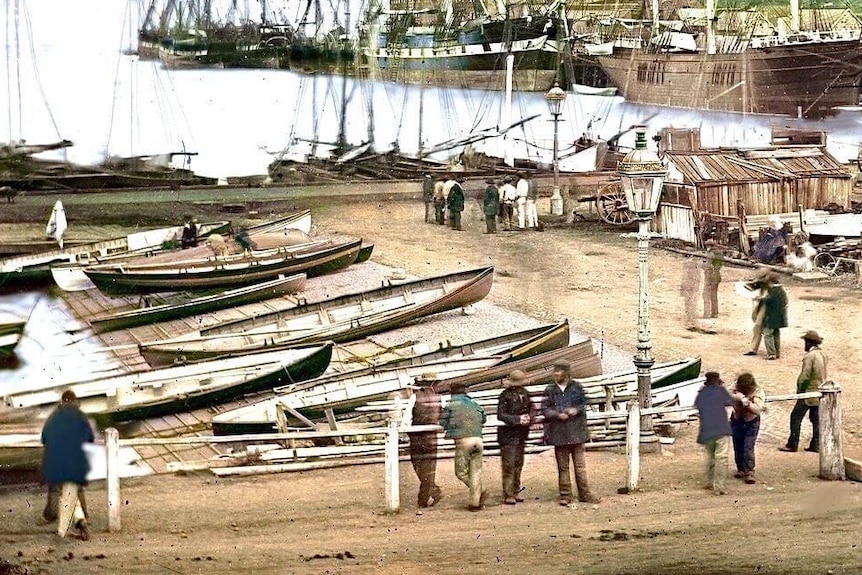 People stand around small wooden boats with the river behind them.