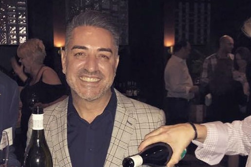 A man smiles while holding a bottle of wine and a glass of wine.