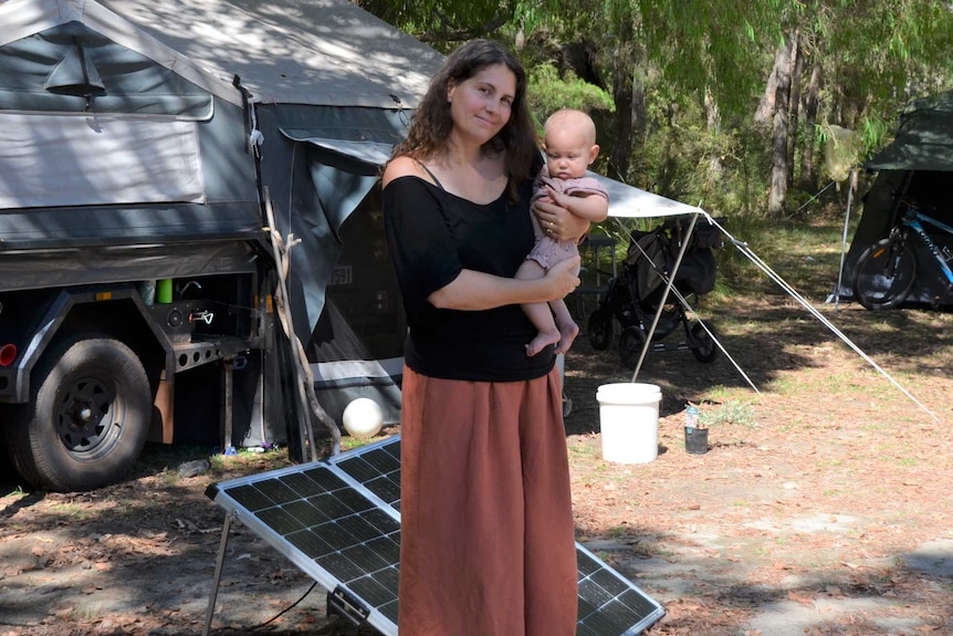 A woman with long dark hair holds a baby while standing in front of a camper trailer and tents.