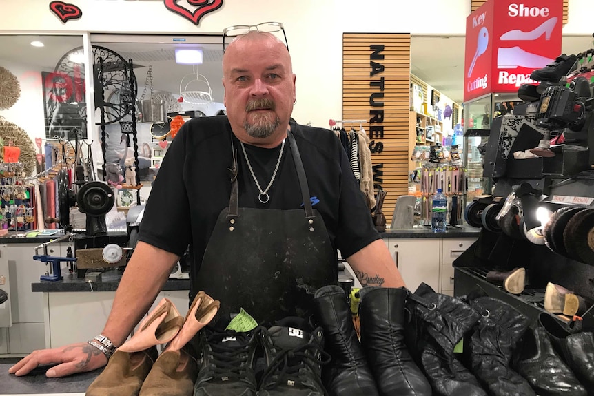 A bald man with glasses on his head stands in front of shoes in a cobblers shop.