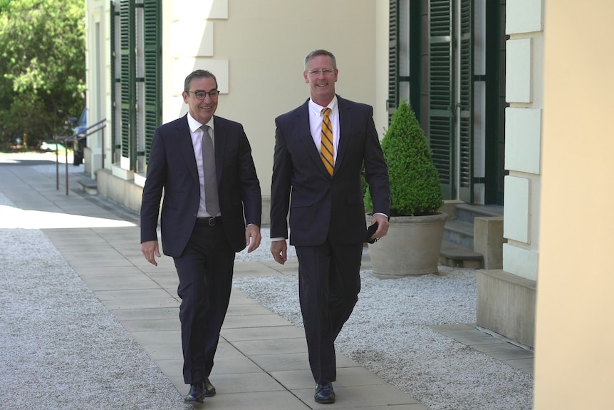Two smiling men wearing dark coloured suits walk on a paved pathway outside government house