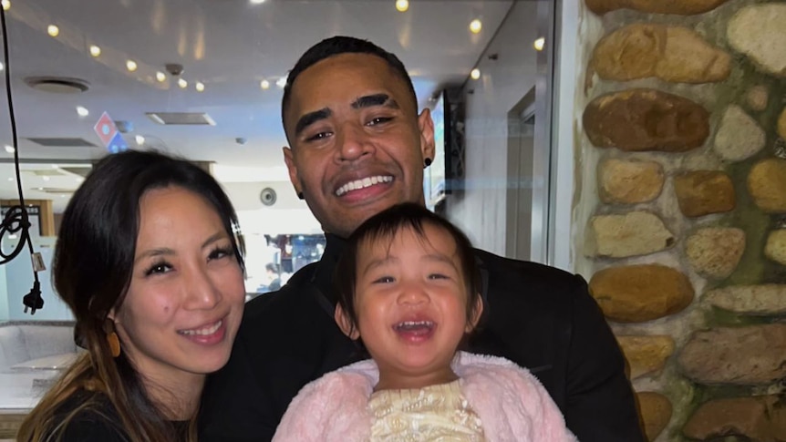 Ray and Rachel Fugawai holding their young daughter, looking and smiling at the camera. 