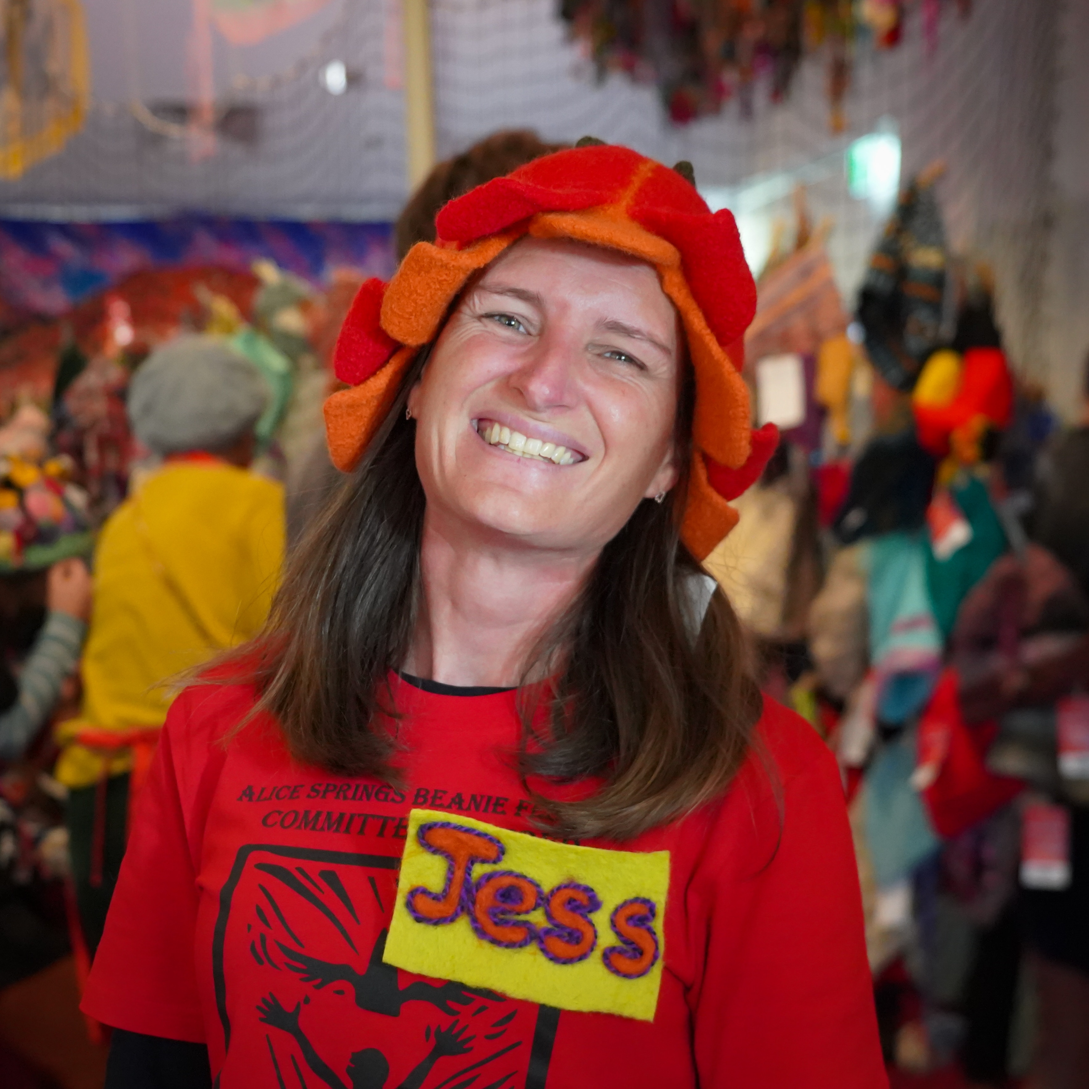 A woman smiling at the camera while wearing a red and orange felt beanie on her head, it is shaped like a blooming flower.