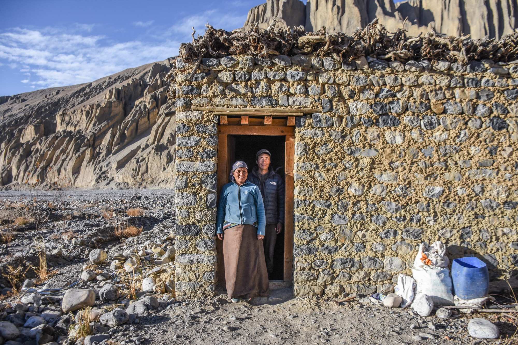 A couple stand in front of a stone home in a mountainous area
