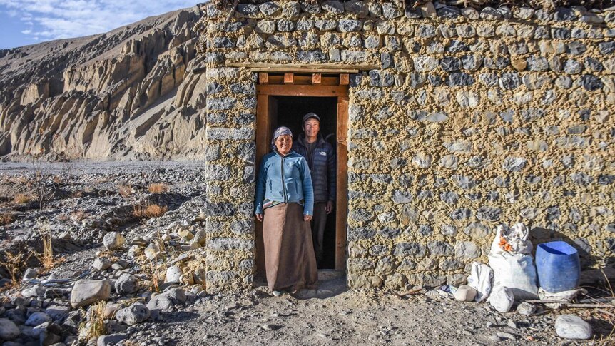 A couple stand in front of a stone home in a mountainous area