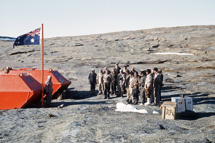 A colour photo from 1954 showing an Australian flag on a wooden pole and a group of expeditioners standing on rocky ground