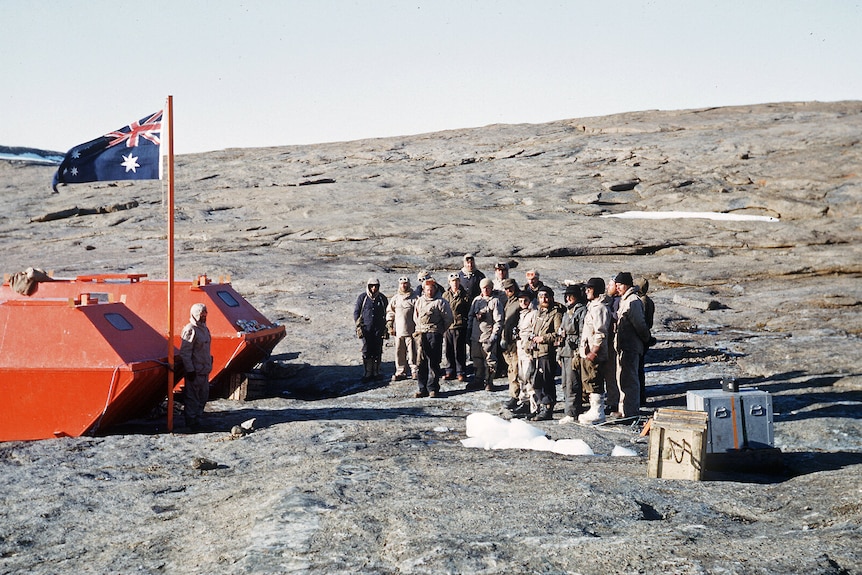 A colour photo from 1954 showing an Australian flag on a wooden pole and a group of expeditioners standing on rocky ground