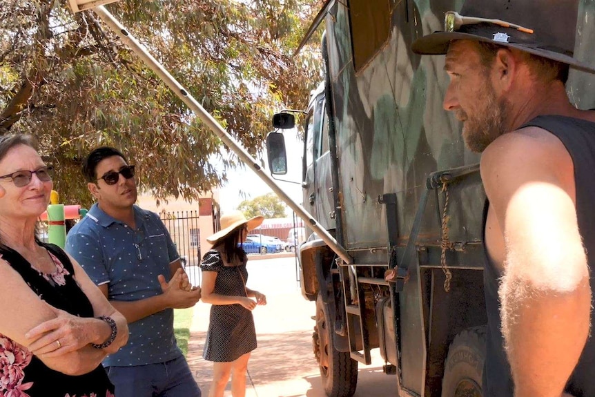 Exhibition visitors listen to a man in a broad-brimmed hat and a singlet as he stands by a truck.