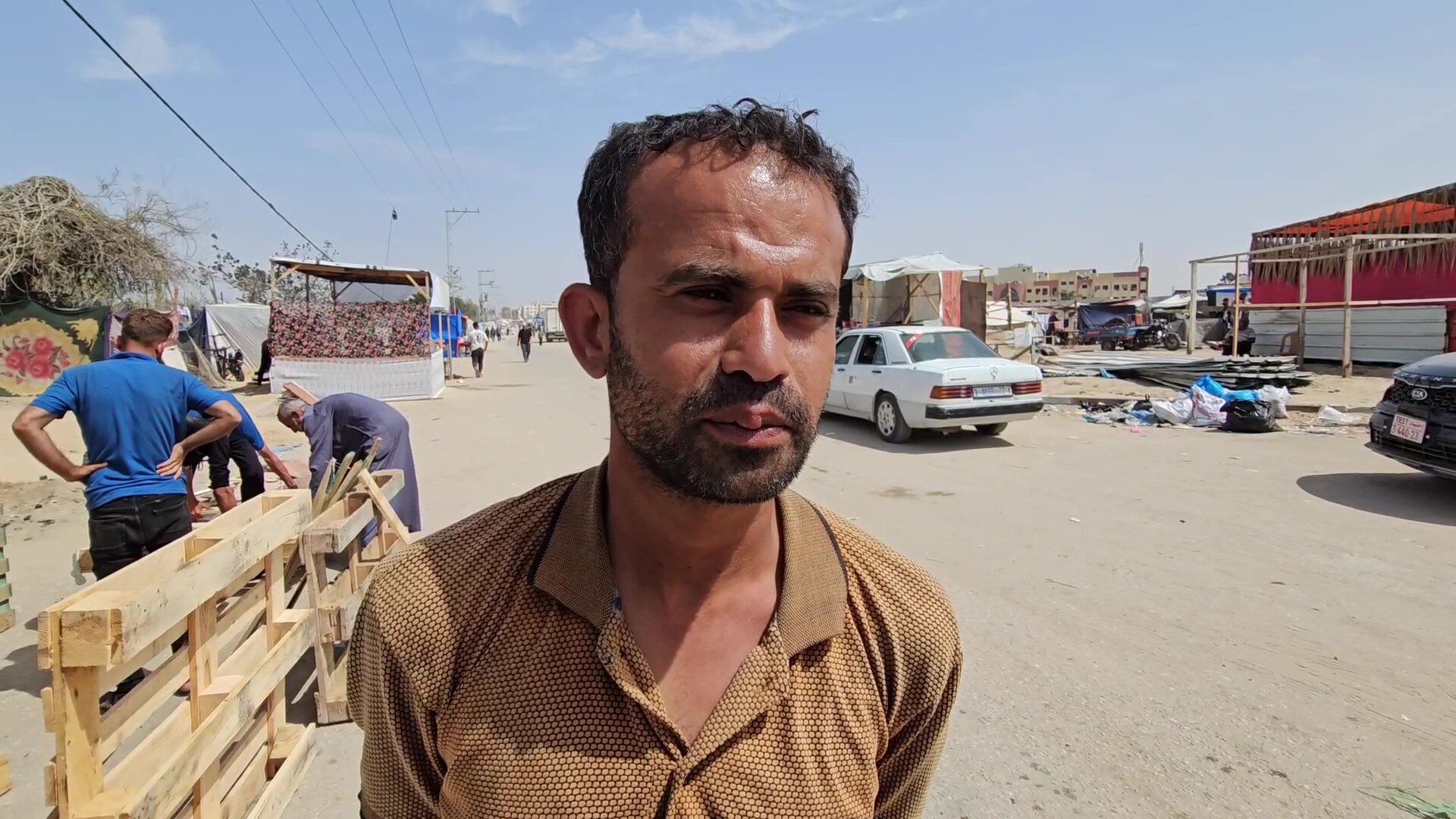 A man in a brown shirt stands outside in the sun.