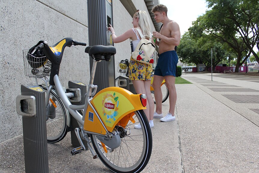 A young man and woman rent a yellow bicycle from a CityCycle terminal at Southbank in Brisbane.