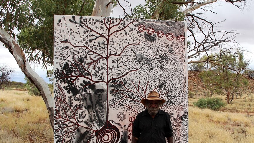 Peter Mungkuri stands beside his painting which is in a tree