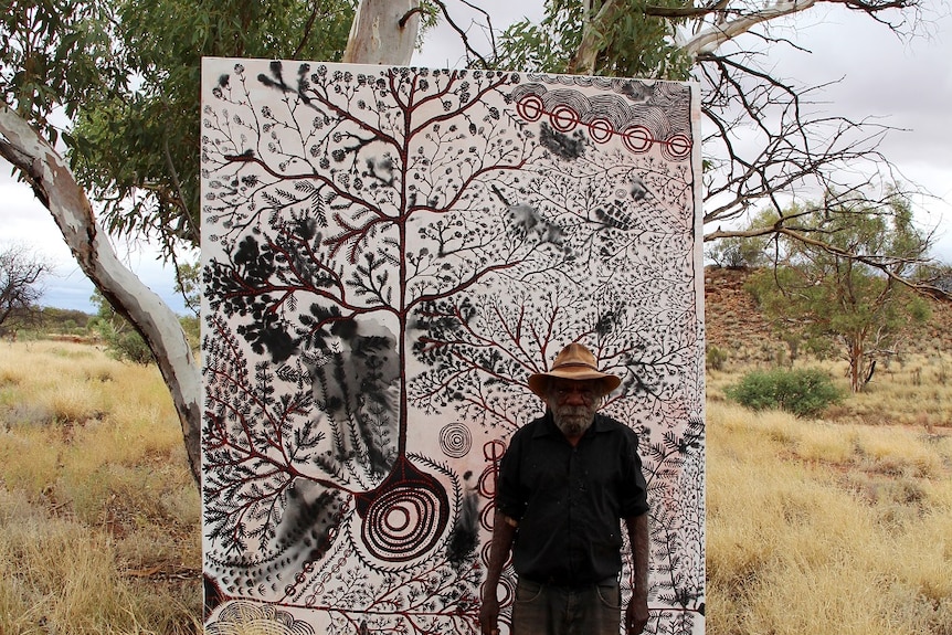 Peter Mungkuri stands beside his painting which is in a tree