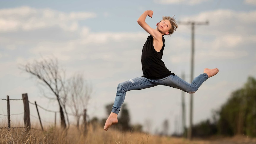 Daley in a split leg pose mid air wearing a black muscle tee and jeans on the side of a rural road.