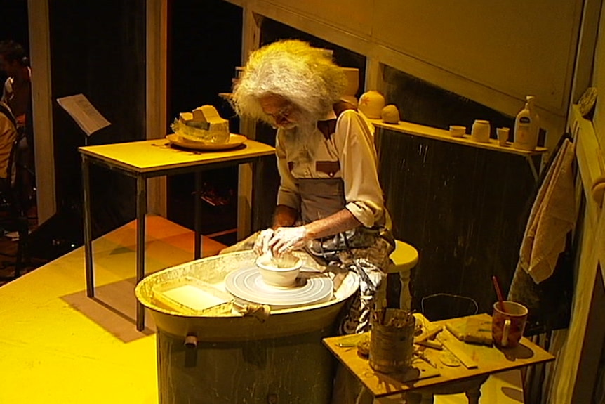 Uncle Jack Charles works at a pottery shelf on stage under yellow lighting.