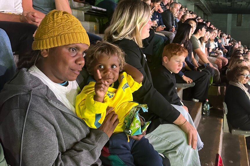 A mother holds her two-year-old son in the stands of a swimming stadium, the son wears an Australian jersey.