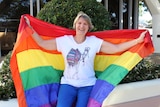 A woman in her 60s sits on a bench, smiling and holding the rainbow flag behind her.