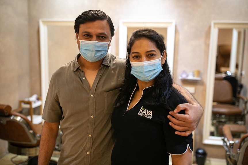 A man and a woman wearing face masks stand in a beauty salon looking at the camera. 