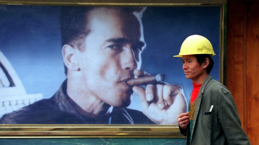 A Chinese man wearing a construction hat walks past a 1990s poster of Arnold Schwarzenegger smoking a cigar