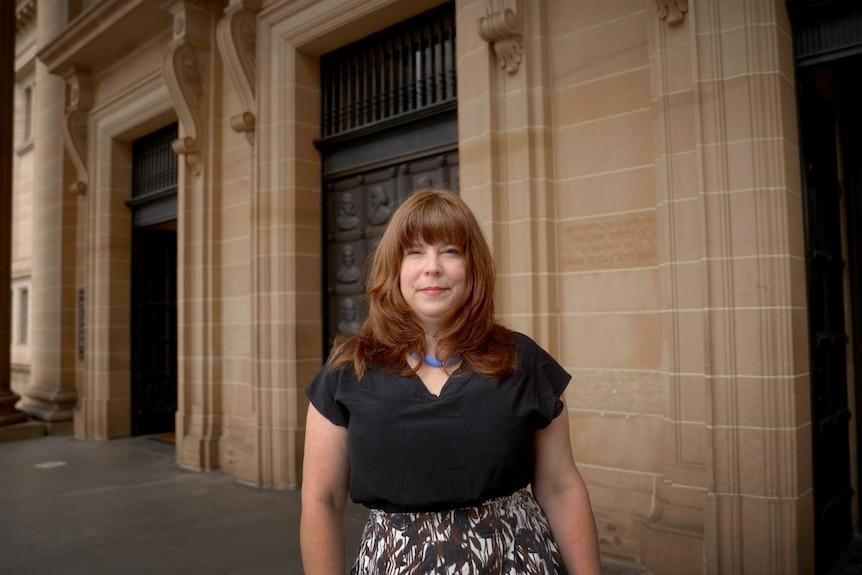 A woman with brown hair and a black top smiles in front of a sandstone building