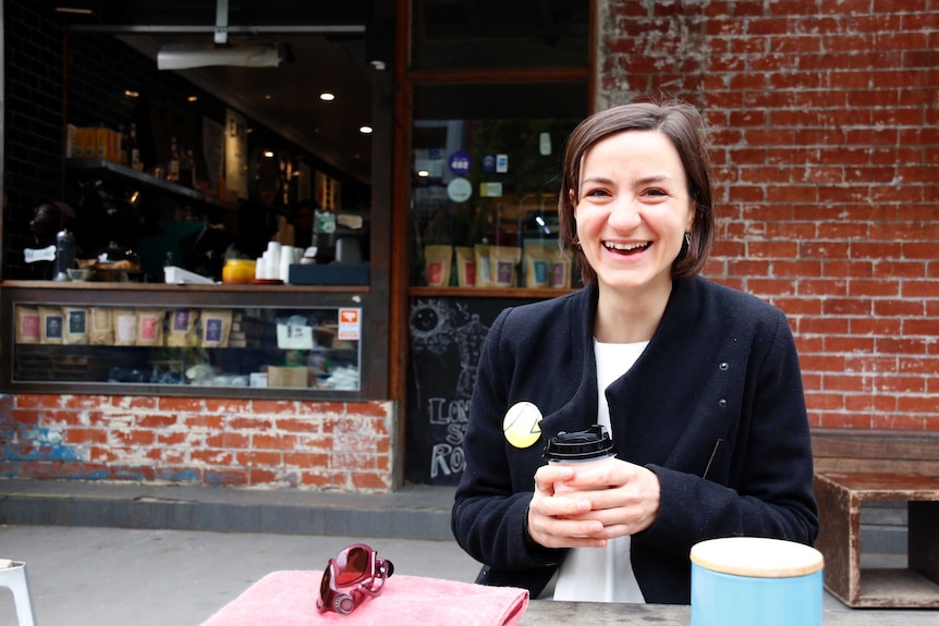 A woman sitting at bench, holding a coffee and laughing.