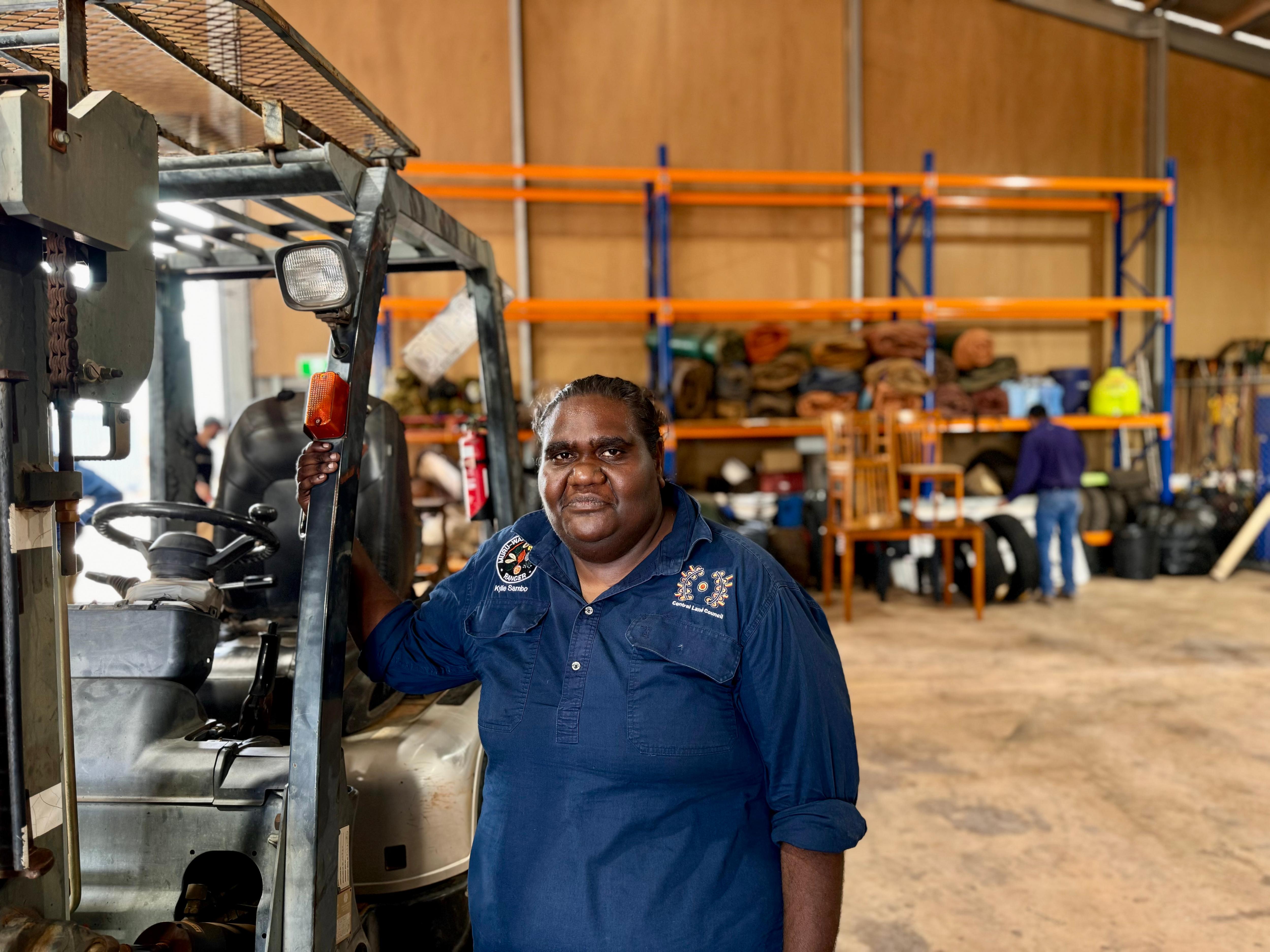 An Aboriginal woman wearing a navy work shirt holds onto the frame of a buggy in a work shed and looks at the camera