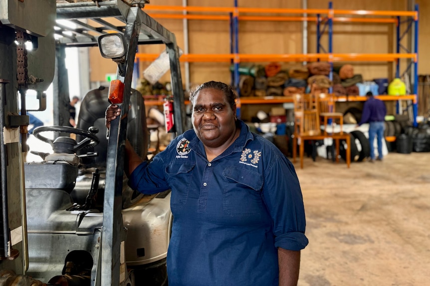 An Aboriginal woman wearing a navy work shirt holds onto the frame of a buggy in a work shed and looks at the camera