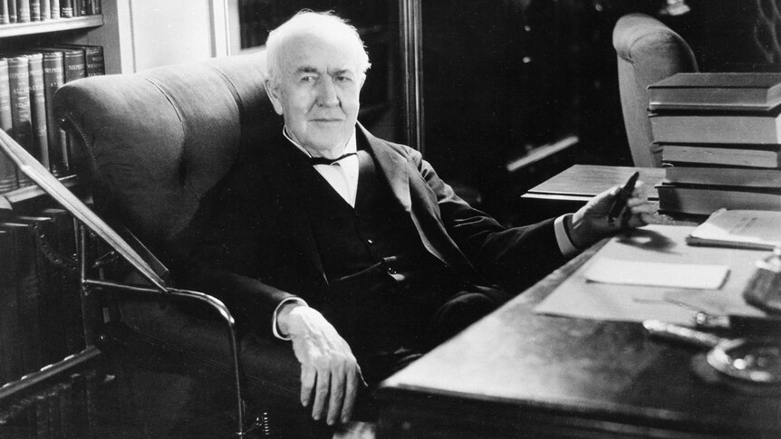 an older edison reclines at his office, hand wresting on the desk