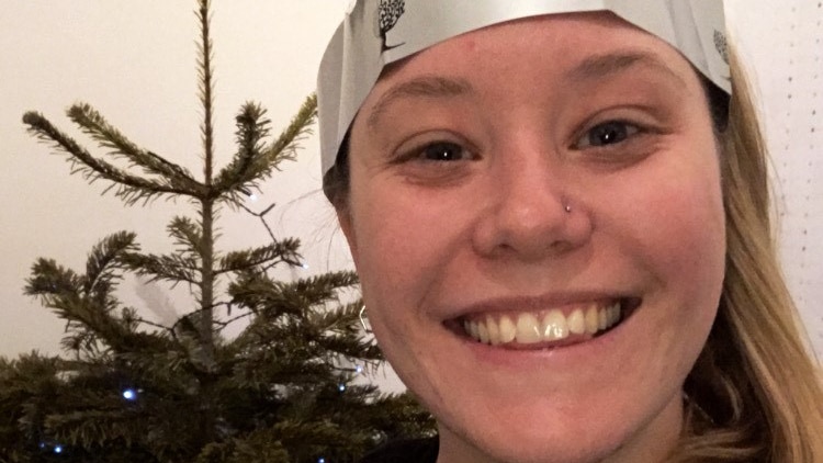 Emily Regan stands in front of a Christmas tree smiling. She is wearing a party hat.