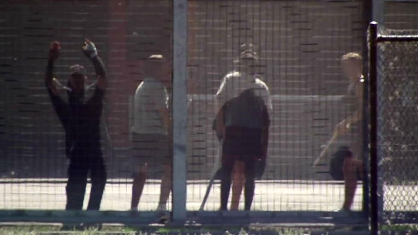 Several detention centre inmates in the yard. One is bashing his hands against the fence.