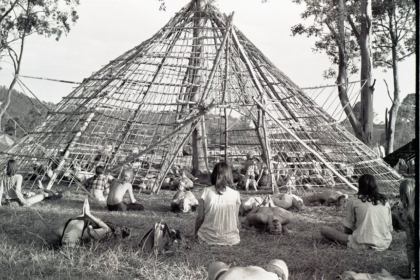 A black and white photo of people outside sitting around a structure made of wood and sticks
