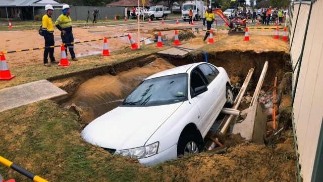 emergency workers stand around a hole in a street verge. A whole sedan car is in the hole.