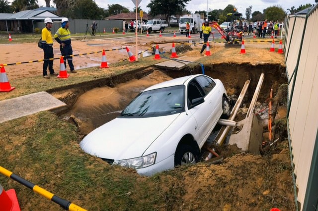 emergency workers stand around a hole in a street verge. A whole sedan car is in the hole.