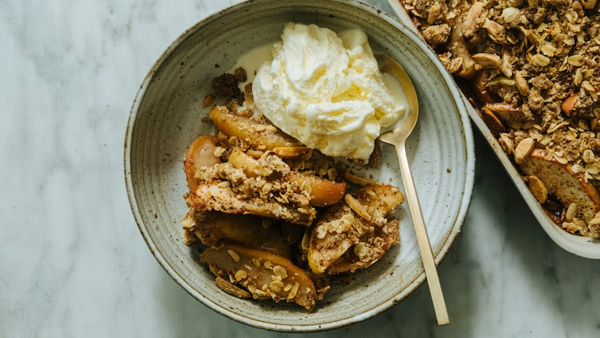 A bowl of apple crisp with a scoop of vanilla ice cream and a spoon. A baking tray of apple crisp sits behind it.
