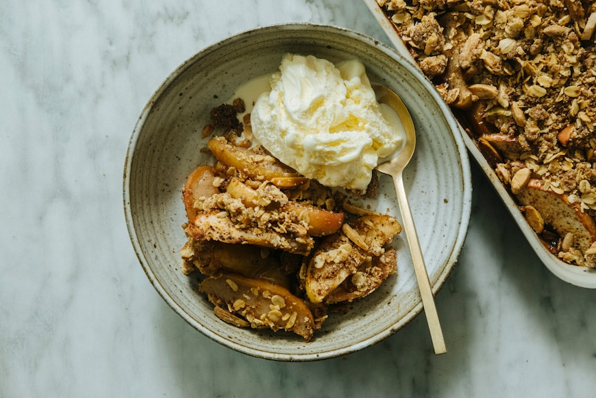 A bowl of apple crisp with a scoop of vanilla ice cream and a spoon. A baking tray of apple crisp sits behind it.