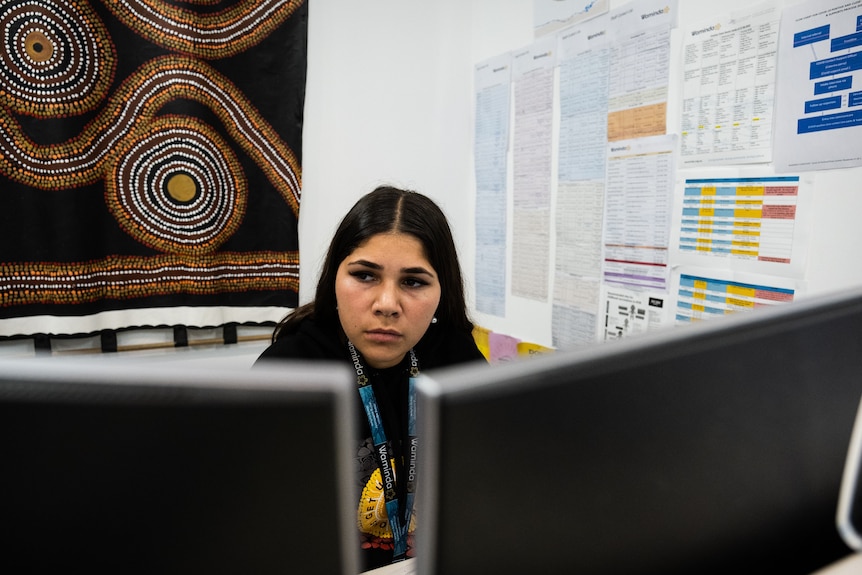 A woman sits at her computer desk. There is an Aboriginal artwork on the wall behind her. 