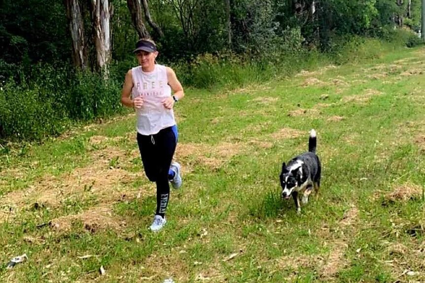 a woman running running in park, white top, black and blue tights, sun visor, black and white dog running beside her.