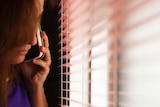 A woman uses her mobile phone while looking through horizontal blinds.