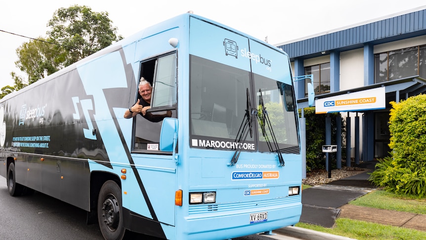 A man hangs sticks his head out the side window of a blue and black bus with his thumbs up. 