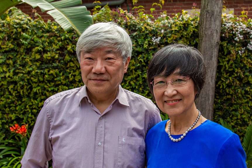 A man with white hair wearing a checked shirt stands next to a woman with short black hair in a blue top.