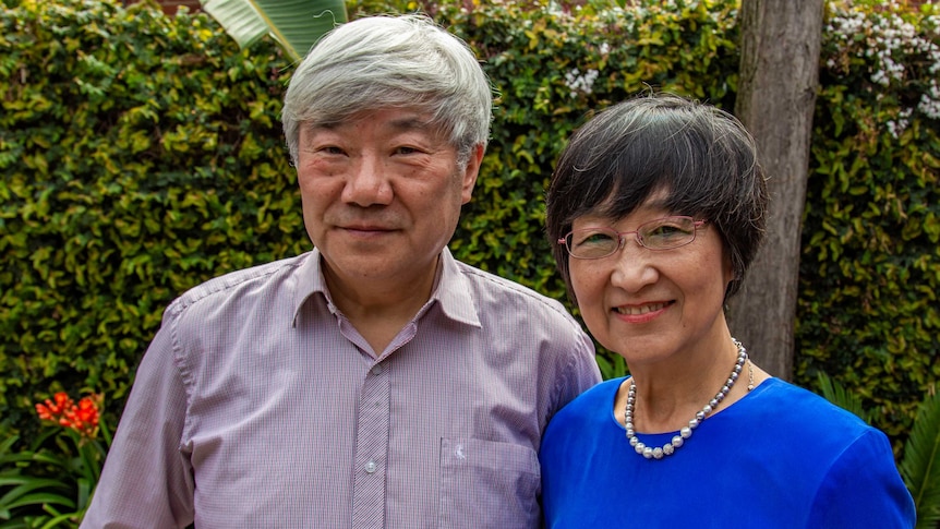 A man with white hair wearing a checked shirt stands next to a woman with short black hair in a blue top.